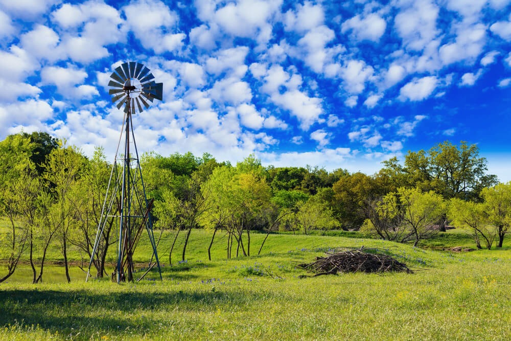 A ranch windmill in a green Texas pasture under a blue sky full of small white clouds